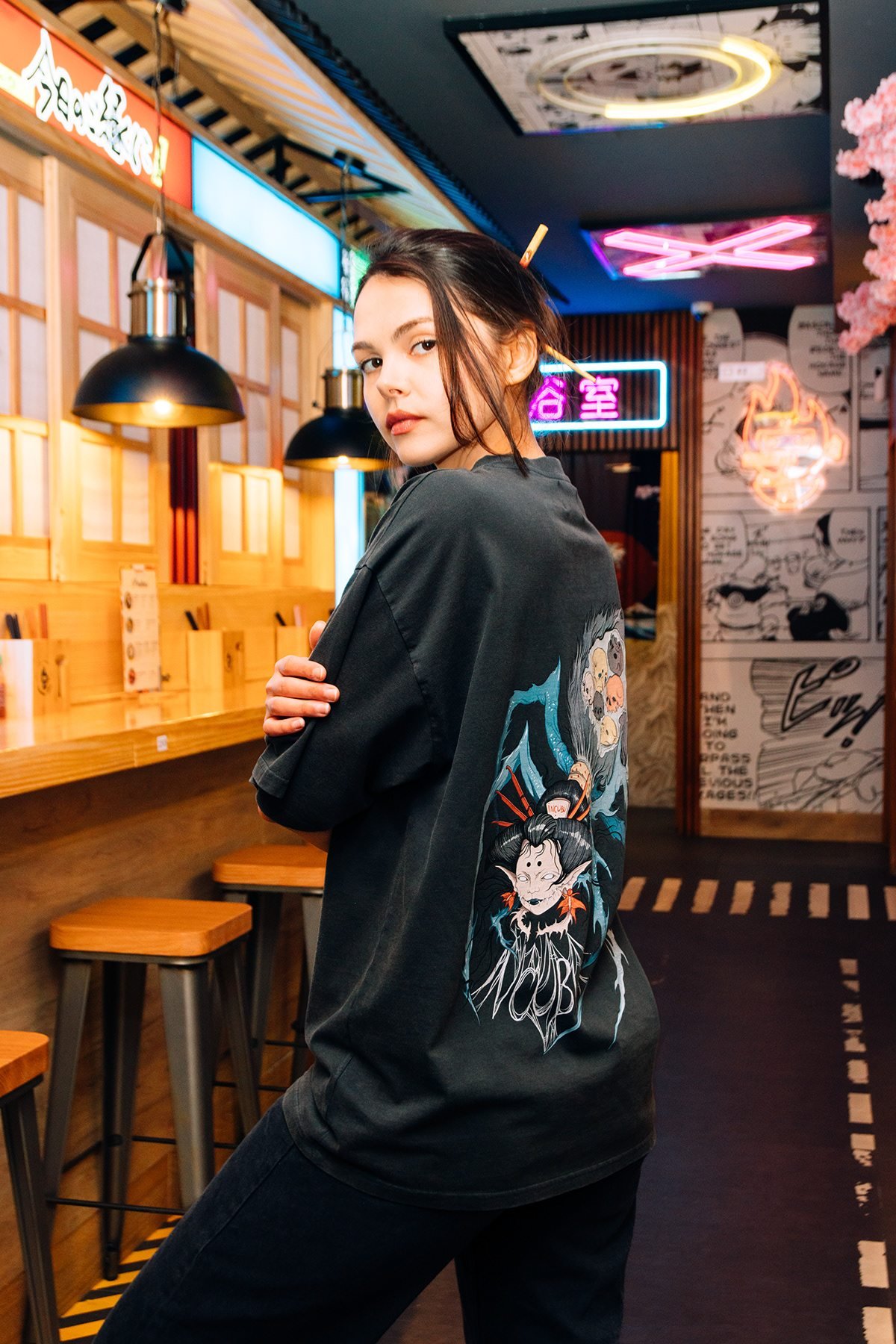 Mujer con camiseta negra estampada mira a cámara junto a una barra de local con luces de neón.