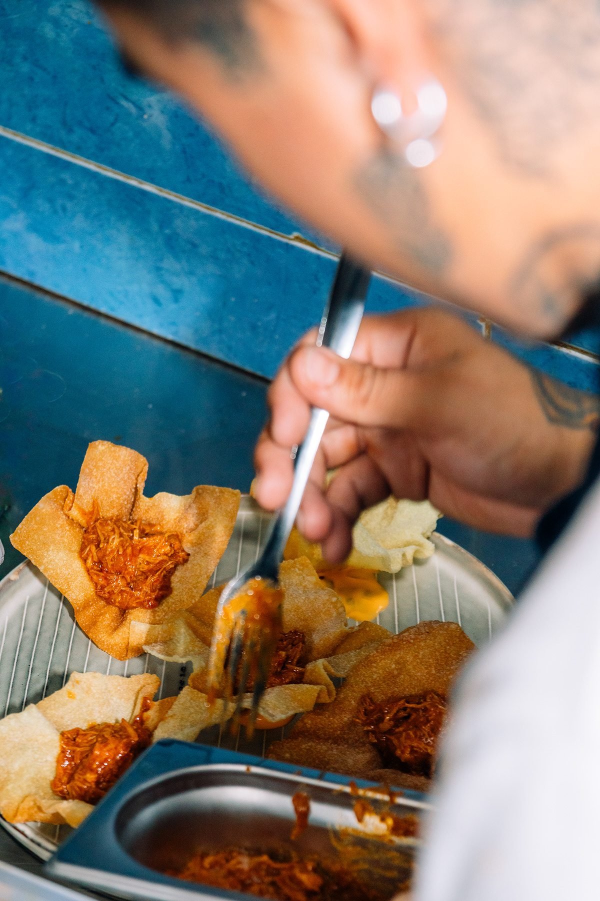 Persona con tatuajes sirve con un tenedor comida con salsa roja en una bandeja metálica.