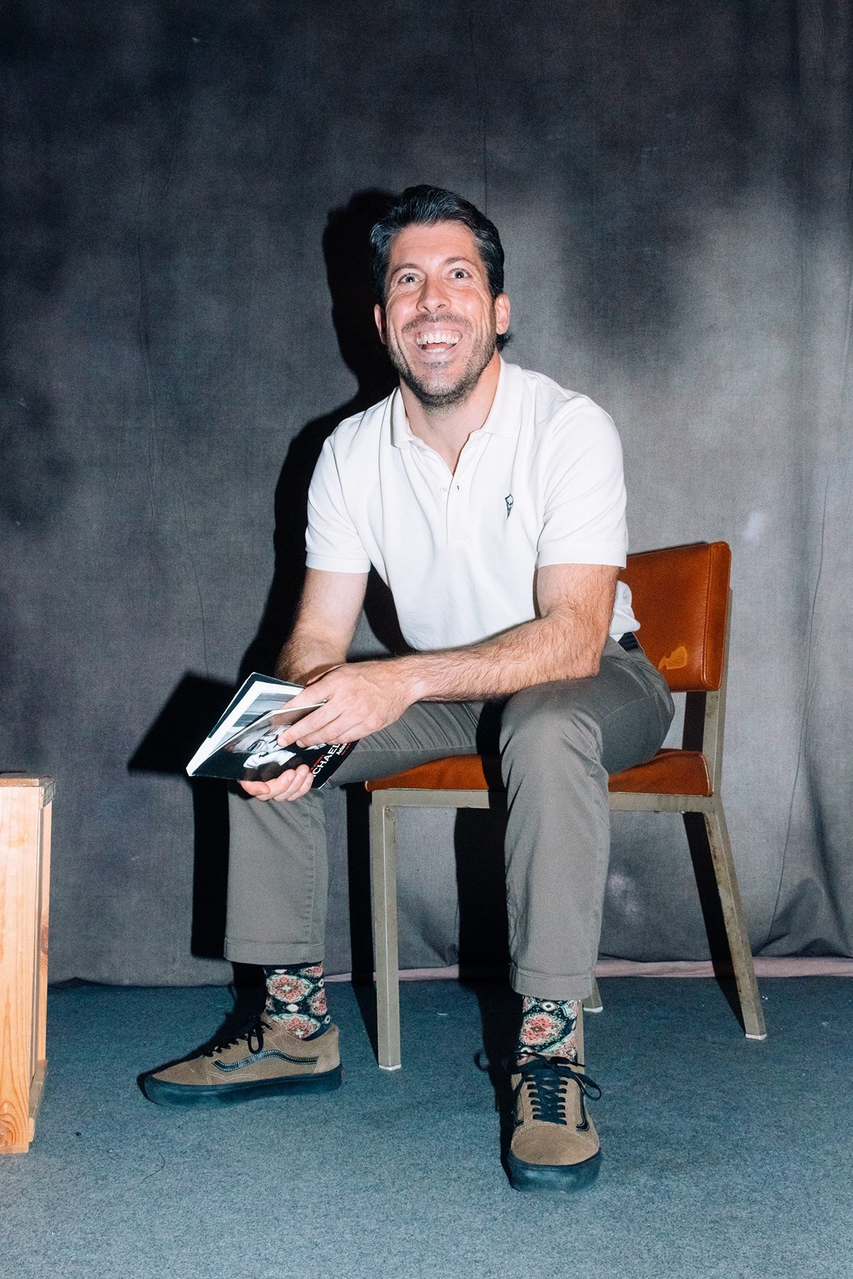 Hombre con polo blanco sentado en una silla sobre fondo gris, con un libro en la mano.